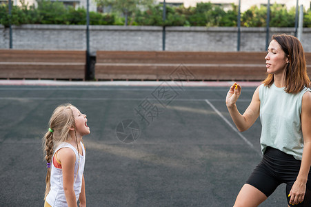 妈妈和女儿在户外运动场上玩得开心 一个高加索女人扔糖果 一个女孩用嘴捡到糖母亲育儿女士孩子婴儿行动公园成人父母压痛健康高清图片素材