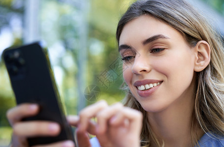 坐在户外的微笑女孩 看着智能手机 使用应用程序在移动声响器上订购美丽的高清图片素材