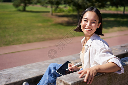 坐在笔上的女孩带着数字平板和图形笔坐在长椅上 笑着微笑 对着镜头转过身来并热烈说话的韩国快乐妇女公园女孩享受药片喜悦绘图自由艺术家黑发女性背景