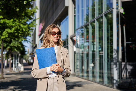 蓝色渐变墨镜戴着墨镜和米色西装的企业女性肖像 拿着蓝色文件夹和办公室文件 去上班 在阳光明媚的日子里走在街上微笑代理人套装职场企业家员工售货背景