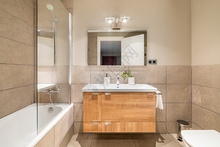 美丽的米色浴室 带木制抽屉的梳妆台台面水槽 浴缸用防溅保护玻璃围起来 正门映在对面的方形大镜子里背景图片