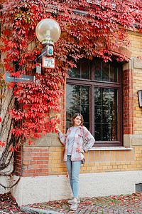 一个美丽的女孩站在欧洲老房子的窗户的背景下 与秋天的飘红交织在一起 旅游与旅游概念 一个年轻女子的漂亮肖像 在秋天秋天的一天在户背景图片