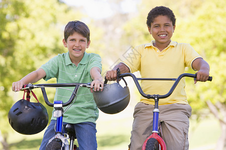骑着铅笔男孩两个男孩在户外的自行车上微笑着笑着公园两个人农村女孩享受安全帽团体男孩们头盔友谊背景