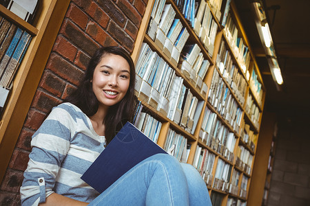 坐在地板上坐在图书馆阅读书墙壁对面的微笑学生大学服装知识教育学校休闲高等教育大学生女性文学背景图片