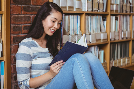 坐在地板上坐在图书馆阅读书墙壁对面的微笑学生女性服装知识快乐教育高等教育文学学校大学生大学背景图片