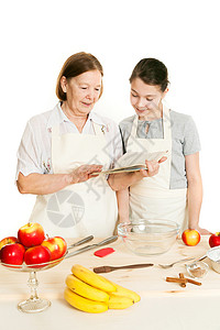 外祖母和孙女读食谱的秘方书厨房女性专家衣服烘烤女孩食物红色阅读糕点背景图片