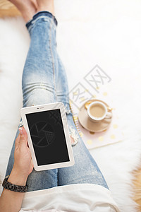 一位穿着破旧牛仔裤的年轻女子坐在家里白色毛皮地毯上的木地板上 手里拿着一台平板电脑 一杯咖啡 笔记本 金色明亮的女性主题在线的高清图片素材
