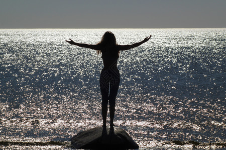 反击一个剪影女孩站在石头上 双臂分开的照片 在海边 在海面上波光粼粼的阳光下海滩海洋喜悦强光天空太阳天堂享受乐趣活力背景