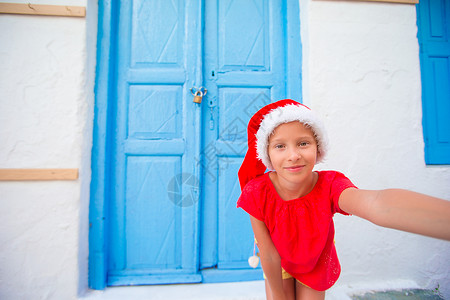 米克诺斯岛Santa Hat的可爱小女孩在希腊村狭窄街道上自拍背景