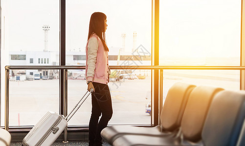 机场贵宾休息室旅行生活方式的概念 出发前在登机口航站楼等候的旅行女游客带着行李在机场窗口看着日出的飞机进行早班飞行背景