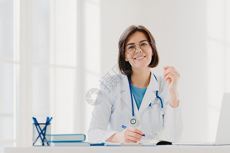 年轻女医生在诊所写纸 为病人开处方 在医院做文书工作 穿着白色医用长袍 坐在舒适的现代办公室里 人 职业 工作概念白色的高清图片素材