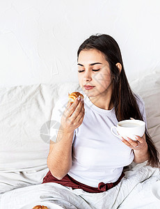 年轻的黑发年轻女人坐在床上吃着羊角面包早餐热情成人女性幸福咖啡气球卧室乐趣甜点生活方式高清图片素材