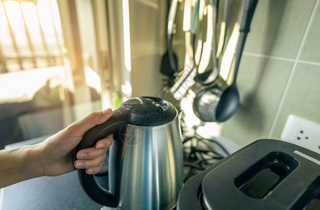 厨房咖啡女人手拿着公寓厨房里电热水壶的把手 亚洲女性早上用银色不锈钢水壶手工准备开水 用于早餐茶或咖啡 厨房用具背景