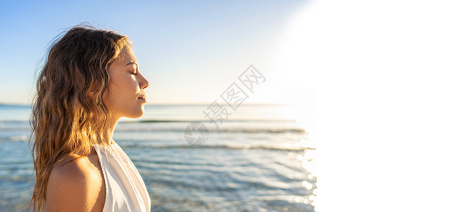 年轻美丽的白人金发女子的侧面肖像 闭着眼睛 穿着白色波西米亚风连衣裙 在日落或黎明时分在海滩上被阳光亲吻 背景是大海 白色复制空健康高清图片素材