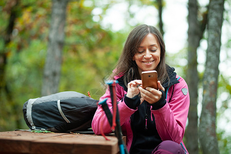 粉色电话皮革女孩躺在森林里的长椅上 背包机里拿着粉红色夹克的手机细胞两极树木远足者运动信号接待远足旅行者技术背景