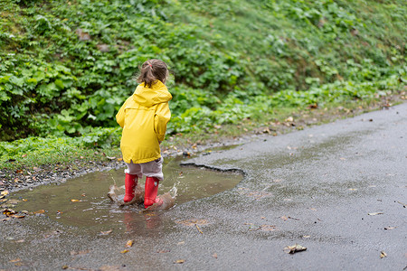 雨坑玩耍的女孩穿着黄色雨衣 同时在降雨时跳上水坑天气季节淋浴衣服孩子快乐乐趣幸福外套雨滴背景