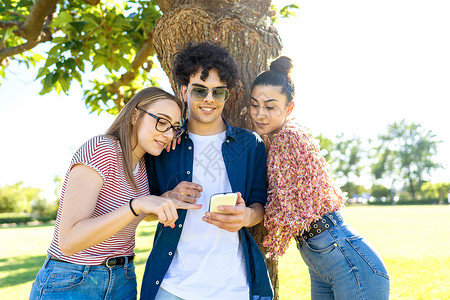 三名学生在公园里使用智能手机指指点点并在显示屏上发短信 玩得很开心 两个漂亮的女孩和她们最好的男朋友在社交网络上分享快乐的假期时外部高清图片素材
