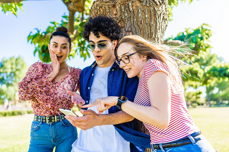 城市公园里的三个年轻朋友惊讶地指着智能手机 享受移动手机上的社交网络 年轻人在大自然中与 Wi-Fi 互联网连接技术一起玩乐移动的高清图片素材