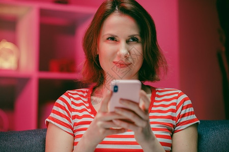 年轻女性拥有智能电话 透过手机屏幕笑笑使用移动工具在社会媒体聊天中购物玩乐游戏 坐在家中的库奇Couch上连接信使房间技术娱乐房看比赛高清图片素材
