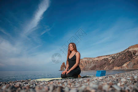 长头发的年轻女性 穿着黑色运动服紧身裤和上衣的健身教练 在普拉提前伸展 在阳光明媚的日子里在海边的瑜伽垫上 女性健身瑜伽常规概念绑腿高清图片素材