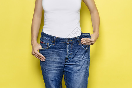 饮食的概念 适当的营养 减肥 展示宽松牛仔裤和她减肥的苗条女人 黄色背景中突显的女式白色 T 恤和超大号牛仔裤身体衣服尺寸活动横背景图片