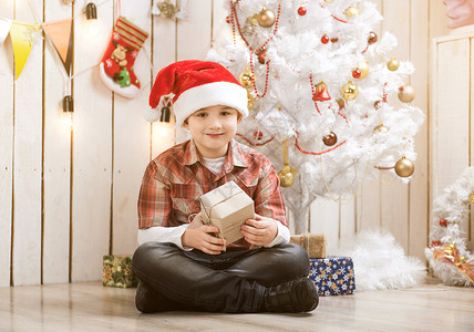 圣诞节帽子男孩坐在新年树旁边的地板上背景