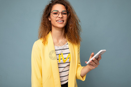 迷人的年轻女性的画像 一头卷曲的深金色头发 身穿黄色夹克 戴着光学眼镜 在背景中被隔离 拿着手机看着相机背景图片