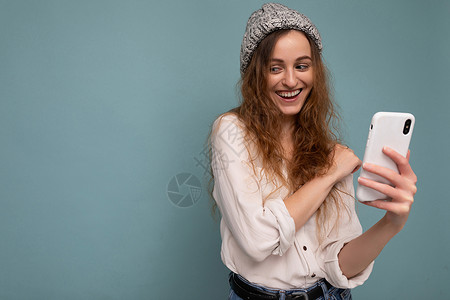 蓝色背景墙上隔着蓝色背景墙 身穿休闲白衬衫和灰色帽子 手持电话 使用在线交流的漂亮年轻卷发女性的照片背景图片