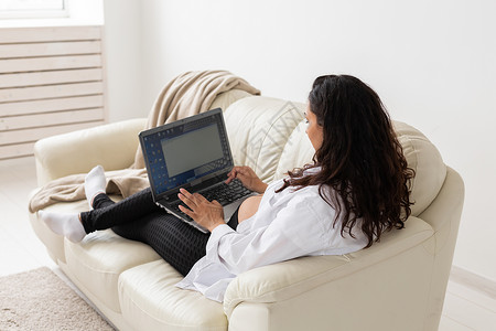 孕妇在家中客厅坐在沙发上时使用膝上型笔记本电脑母亲黑发母性公寓怀孕长椅婴儿阅读女性药片手高清图片素材