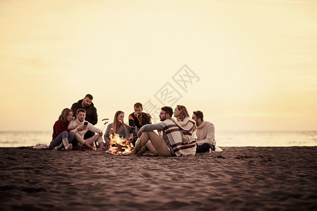 坐在海滩火炉边的年轻朋友团体温暖野餐篝火朋友们友谊香肠青年火焰细胞烧烤背景图片