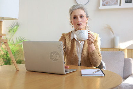 快乐的中年妇女在家里用笔记本电脑喝咖啡浏览高清图片素材