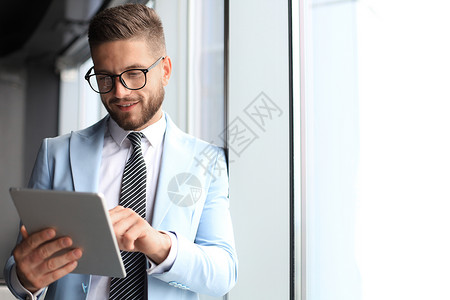 现代正装商务人士在办公室靠近窗口时使用数字平板电脑 同时站在办公室窗户附近职业阶层眼镜经理数位板商务推销技术衬衫工作背景图片