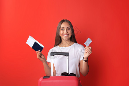 穿暑期散装的年轻旅游女孩 带着太阳镜 红色手提箱 在红色背景下被孤立的护照案件闲暇旅游冒险行李文档手提包游客商业女孩航程高清图片素材