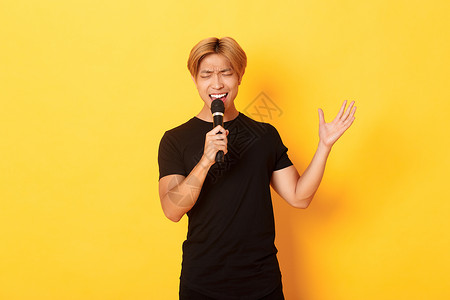 英俊的男性歌手 韩国人唱着歌 在卡拉OK用麦克风 用激情和热情地在黄色背景上站立广告职业唱歌头发金发成人快乐理发工作室促销男人高清图片素材