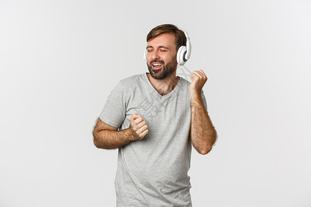 穿着灰色T恤的快乐英俊帅哥肖像 在无线耳机和舞蹈中听音乐 站在白背景上男性成功发型胡须衬衫工作室音乐促销情绪胡子模型高清图片素材