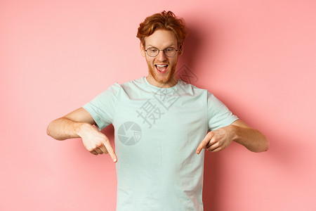 红头发和眼镜的英俊caucasians男子 指着手指 往下看Promo报价 微笑高兴 站在粉红色背景上学生发型成人购物广告粉色红背景图片