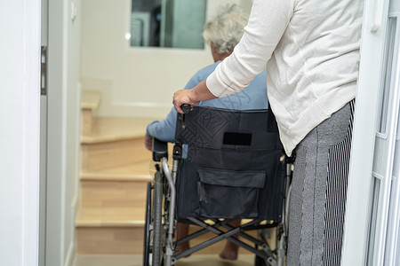 坐在轮椅上的年长或年老的助老妇照顾者扶起家庭楼梯背景