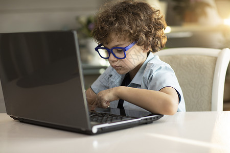 小学媒体素材戴眼镜的卷毛男孩正在用笔记本电脑书 仔细看监视器瞳孔键盘房间专注家庭作业媒体小学头发学习背景