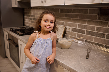 厨房地柜一个快乐可爱的美丽白人小女孩的画像 她穿着蓝色裙子 可爱地微笑着看着镜头 在家里的厨房里准备面团时 她的手臂靠在厨房台面上背景