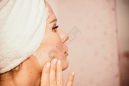 年轻漂亮的女人在洗完澡后使用保湿面膜 头戴毛巾的漂亮迷人女孩站在家庭浴室的镜子前 日常卫生和皮肤护理产品粉刺治疗美容女士皱纹预防调色高清图片素材
