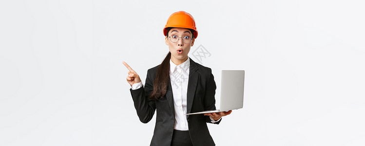 工厂的亚洲女企业家印象深刻 戴着安全帽和西装 手指向左 手持笔记本电脑 在企业展示图表 白色背景商务安全微笑团队员工建筑师项目技背景图片