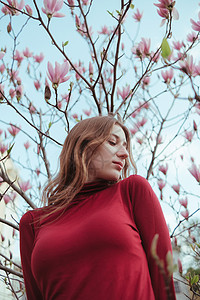 美丽红色木兰花一个穿红衣的黄发女孩 在木兰树的背景之下 与自然团结的概念头发眼睛衬套太阳疗法帽子植物群花朵芳香金发女郎背景