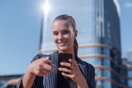 微笑的女士用智能手机指着你城市性格商业城市生活中心街道成功建筑手势女孩背景图片