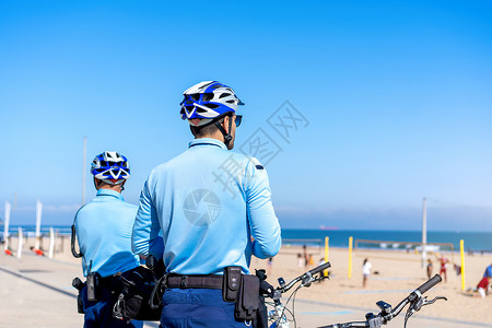 两名警察骑着自行车巡逻海边列车 人们在大西洋海岸的城市公共海滩晒太阳 美国海军陆战队民众热带男人长廊头盔车辆海岸天空街道运输背景图片
