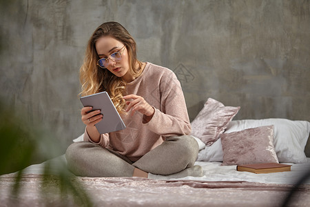 戴眼镜 穿着休闲服的女博主 盘坐在床上 在卧室里用平板电脑工作 学生 博主 灰色墙壁的内部发型高清图片素材