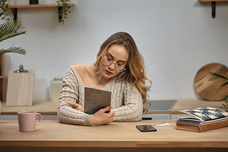 戴眼镜的女博主 米色毛衣 拿着平板电脑 坐在厨房里的木桌旁 上面放着智能手机 塑料卡和杯子 特写在线的高清图片素材