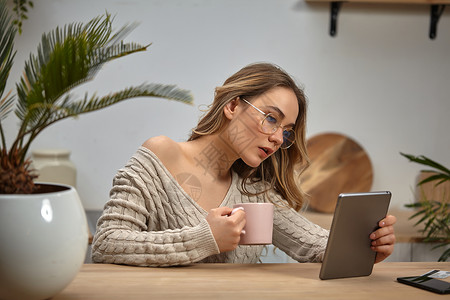 戴眼镜的女博客 美容衣 拿着粉红色杯子和平板电脑 坐在厨房用棕榈 智能手机 塑料卡等木制桌子坐着博主高清图片素材