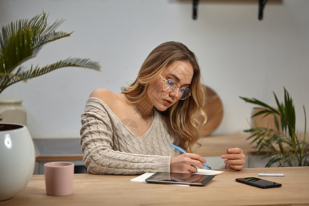 戴眼镜的女人 美容衣 坐在厨房的桌子上 有杯子 棕榈 平板电脑 智能手机和卡片 用笔记本写东西人们高清图片素材
