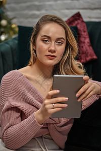 穿着粉红色毛衣的女部落客 坐在灰色沙发上 拿着她的平板电脑 内地装饰着圣诞树年轻的高清图片素材
