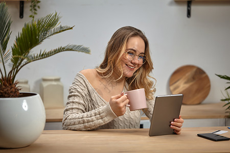 戴眼镜的模特博主 米色毛衣 微笑着 拿着平板电脑 粉红色的杯子 坐在厨房的桌子旁 手掌 智能手机 塑料卡 特写商业高清图片素材